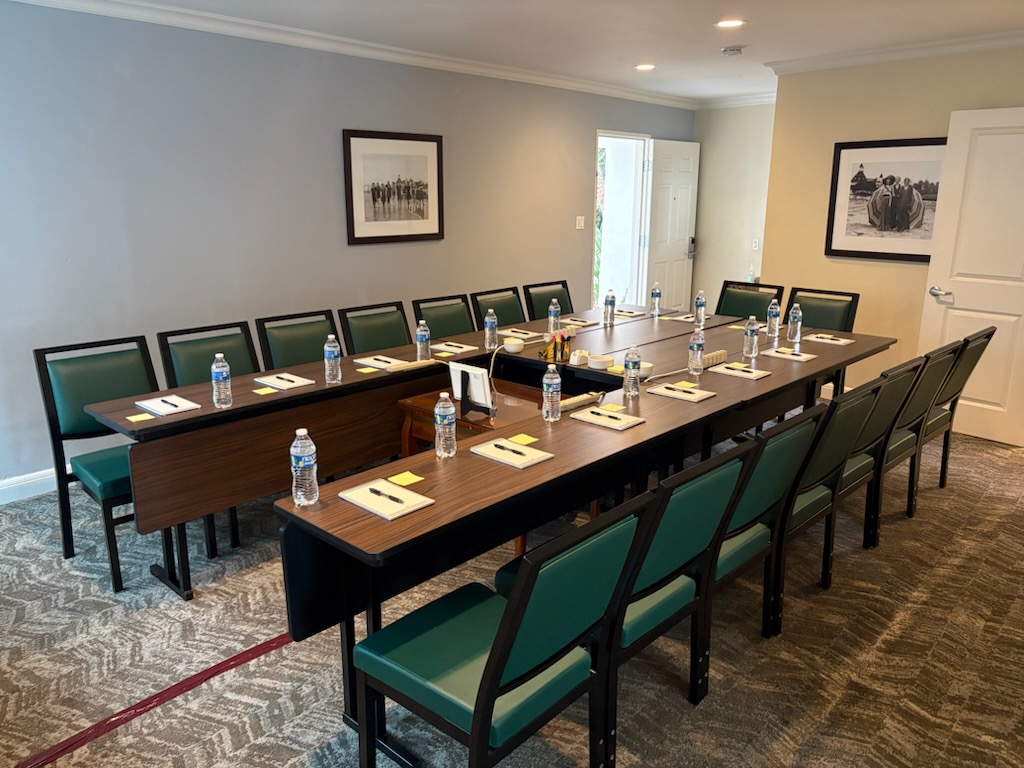 A conference room with two tables arranged in a U-shape, green chairs, notepads, pens, and water bottles at each seat. Framed black-and-white photos hang on the beige walls.