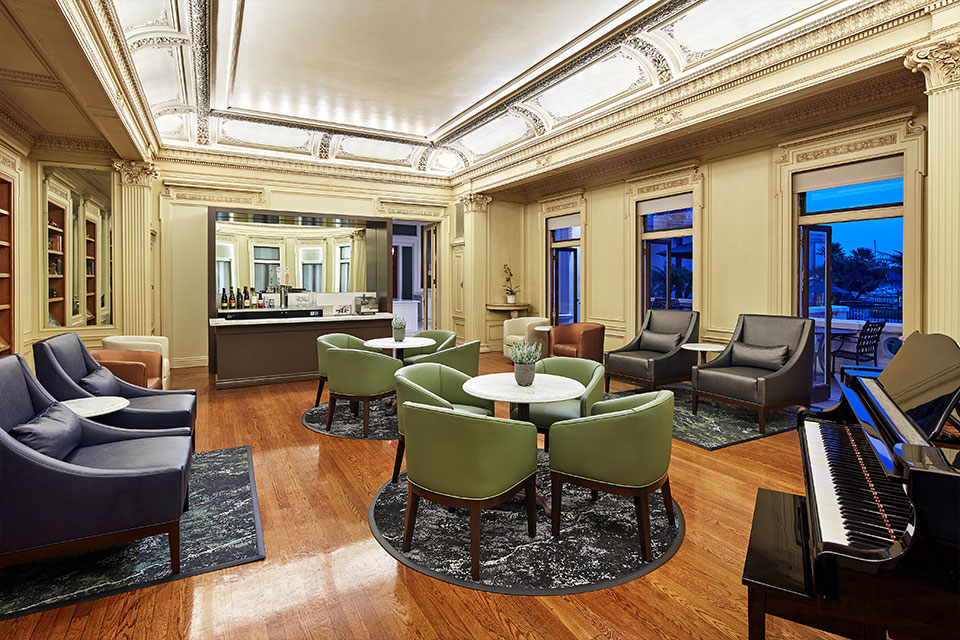 Elegant lounge with green and gray chairs around round tables, a grand piano, wood floors, ornate ceiling, large windows, and a small bar area with wine bottles in the back.