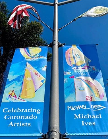 Two colorful banners hang from a pole under blue sky, each showing sailboats on water. The left says Celebrating Coronado Artists. The right reads Michael Ives. An American flag waves above.