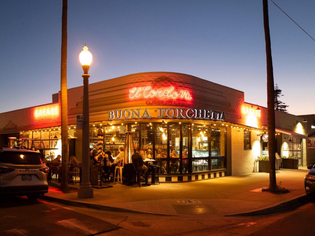 A warmly lit Italian restaurant, Buona Forchetta, with large windows and outdoor seating, is busy with diners at dusk. Neon signs and string lights illuminate the corner building.