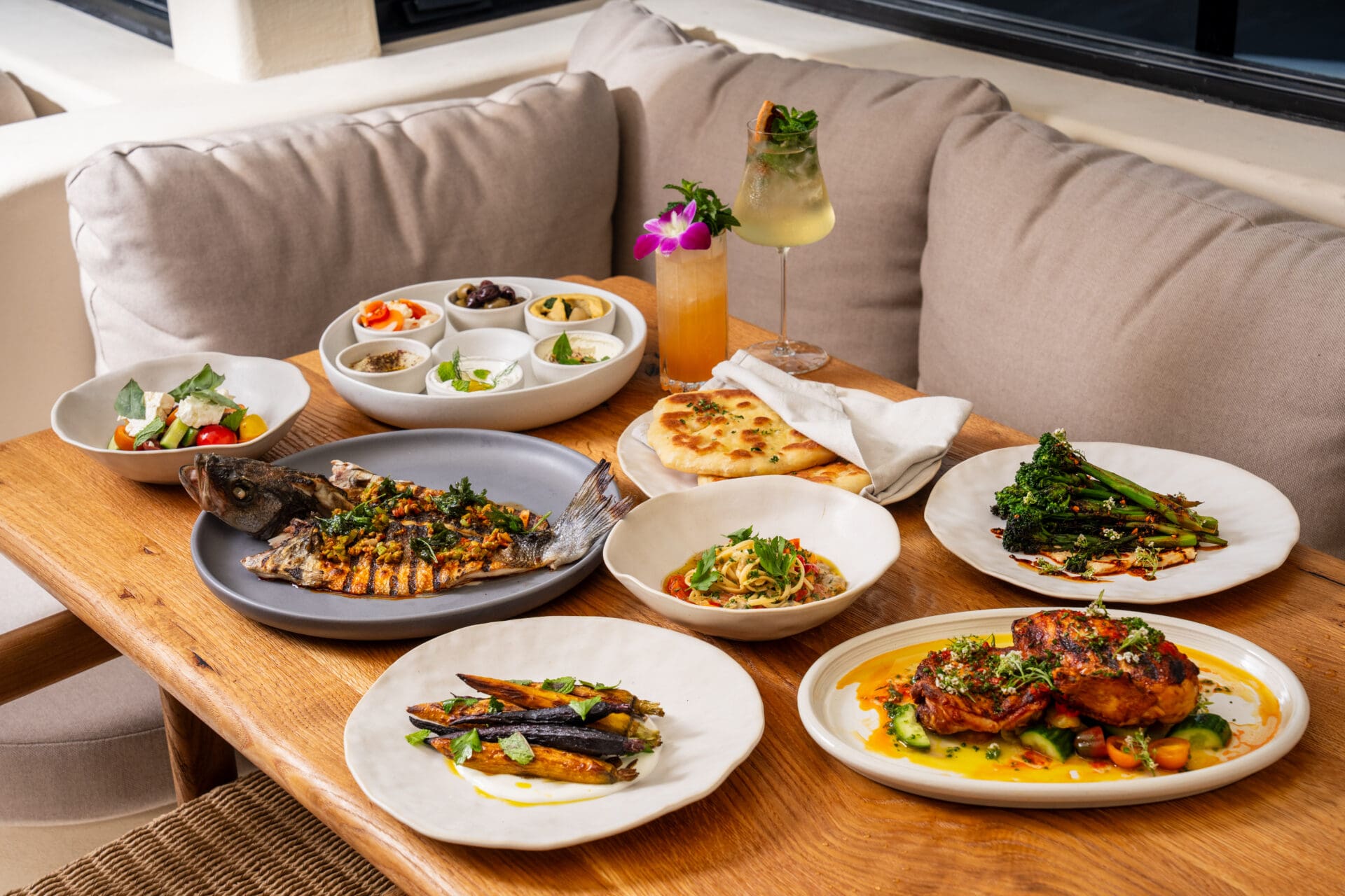 A wooden table set with various Mediterranean dishes, including grilled fish, roasted chicken, vegetables, dips, pita bread, salads, and two cocktails—favorites of many—arranged in a cozy and pet-friendly dining area.