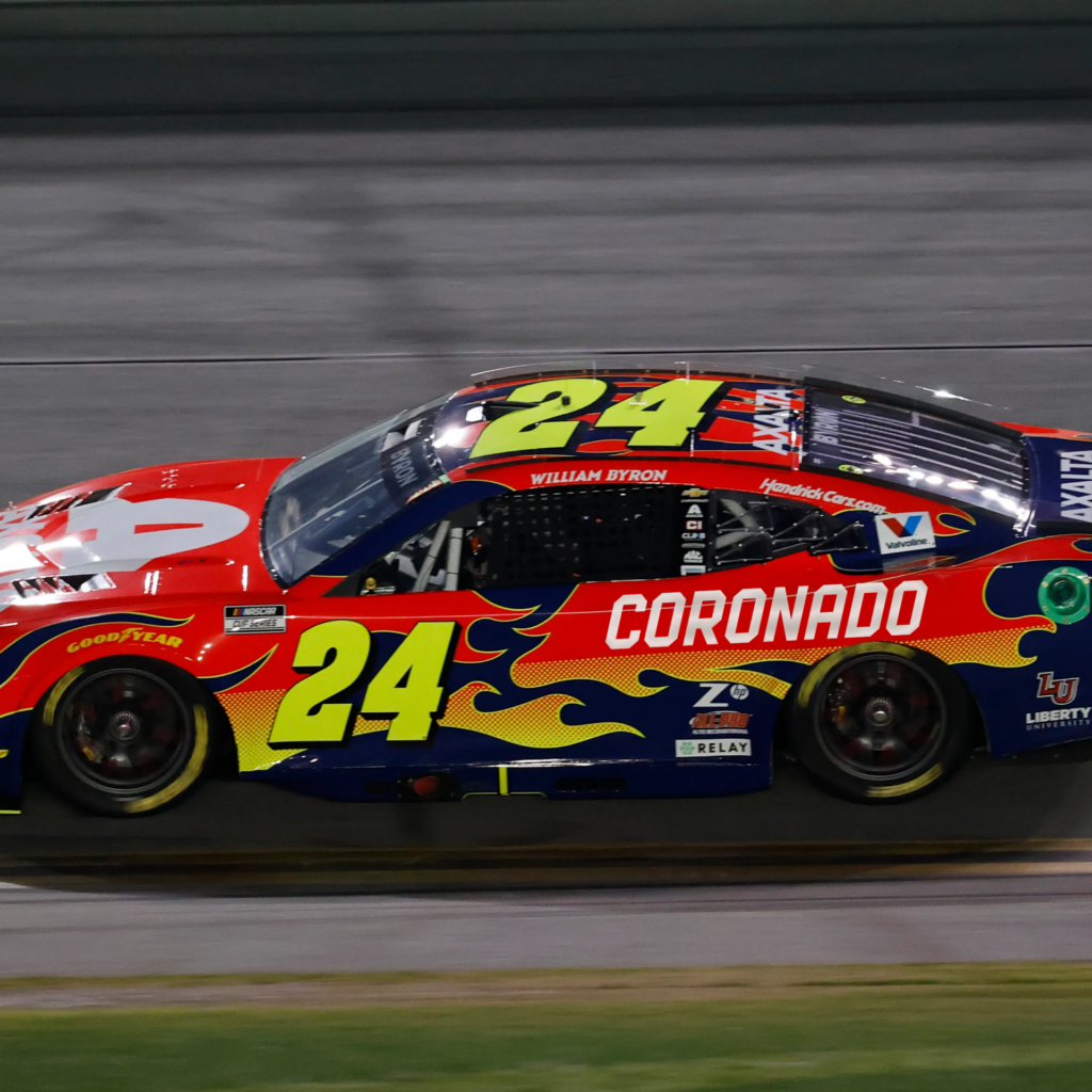 A red and yellow NASCAR race car with the number 24, blue flames, and CORONADO written on the side speeds along a track.