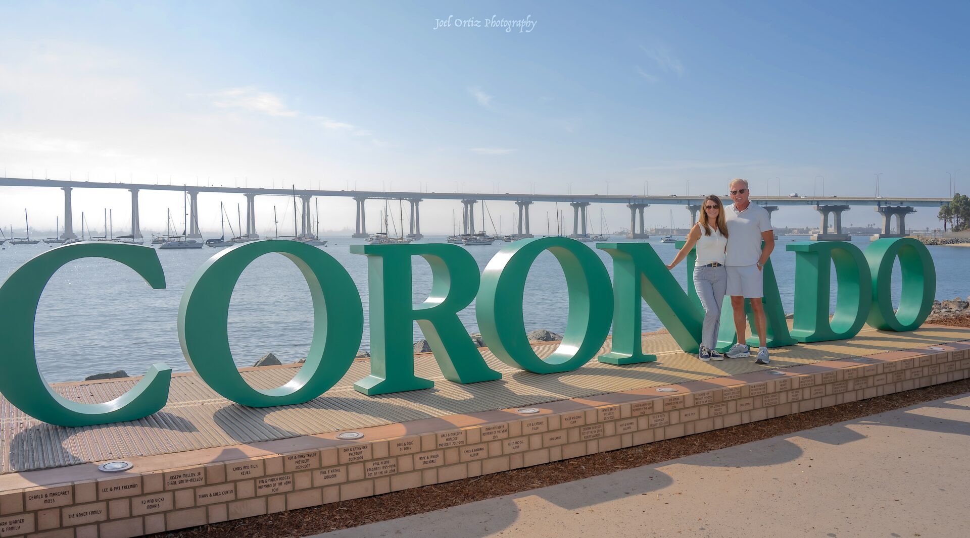 A man and woman stand by large green CORONADO letters near water, with boats and a long bridge in the background under a partly cloudy sky.
