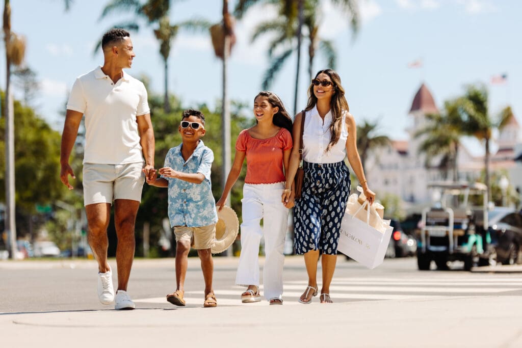 A family of four, dressed in summer clothing, smiles and holds hands while walking outdoors on a sunny day. Palm trees and a historic building are visible in the background. The mother carries shopping bags.