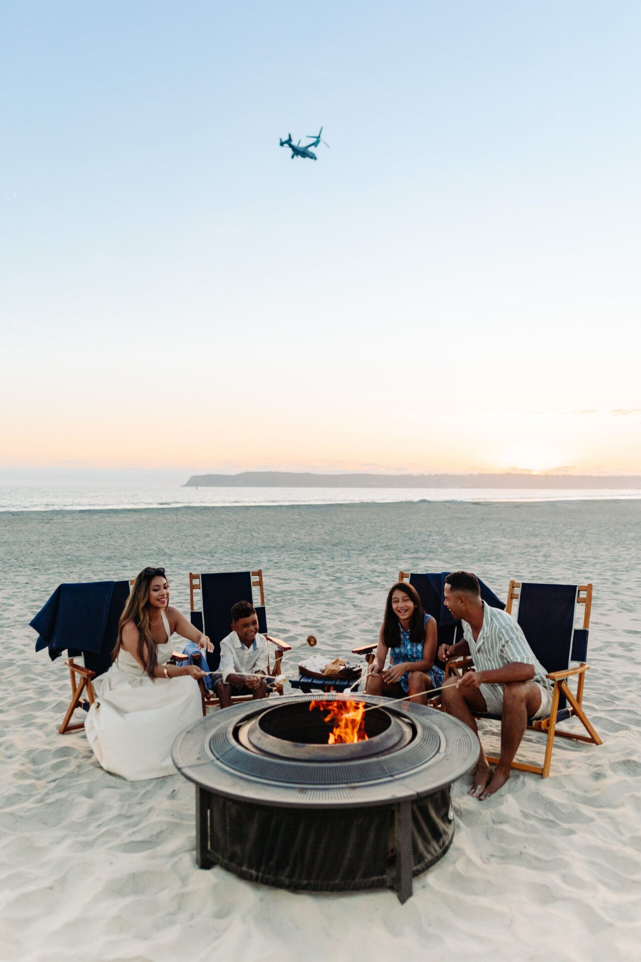 Four people sit around a fire pit on a sandy beach at sunset, roasting marshmallows and smiling. Blue chairs surround them, and an airplane flies overhead in the clear sky.