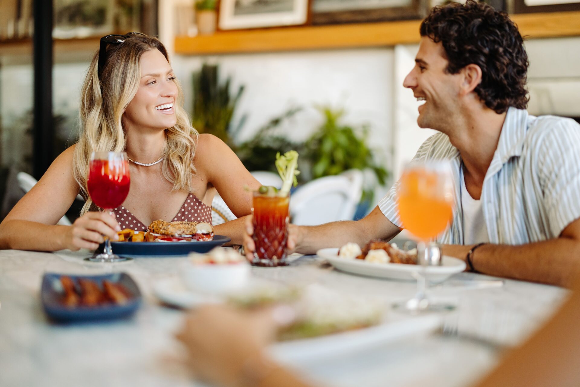 A smiling man and woman sit at a table in a restaurant, holding colorful drinks and enjoying a meal together. Plates of food and various drinks are visible on the table. They appear to be having a cheerful conversation.