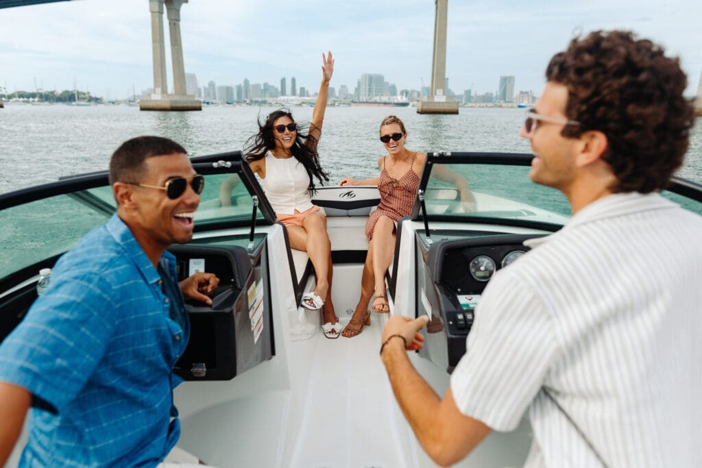 Four friends enjoy a sunny day on a boat. Two women sit at the front, one with her arm raised, while two men stand nearby. All are smiling, wearing sunglasses, with city buildings and a bridge in the background.