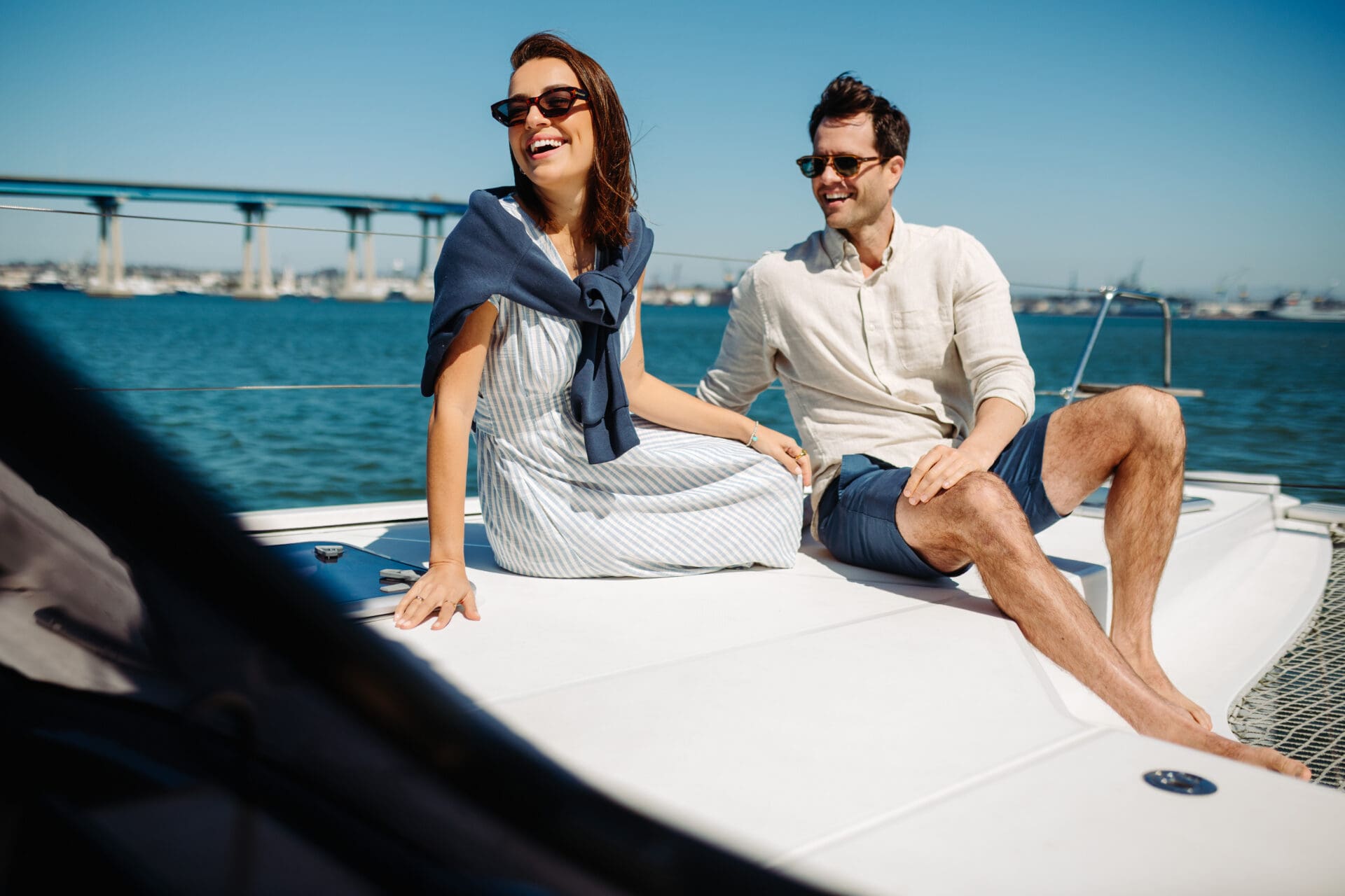 Four friends enjoy a sunny day on a boat, smiling and laughing. Two women sit at the front, one raising her arm, while two men stand nearby. A city skyline and bridge are visible in the background.