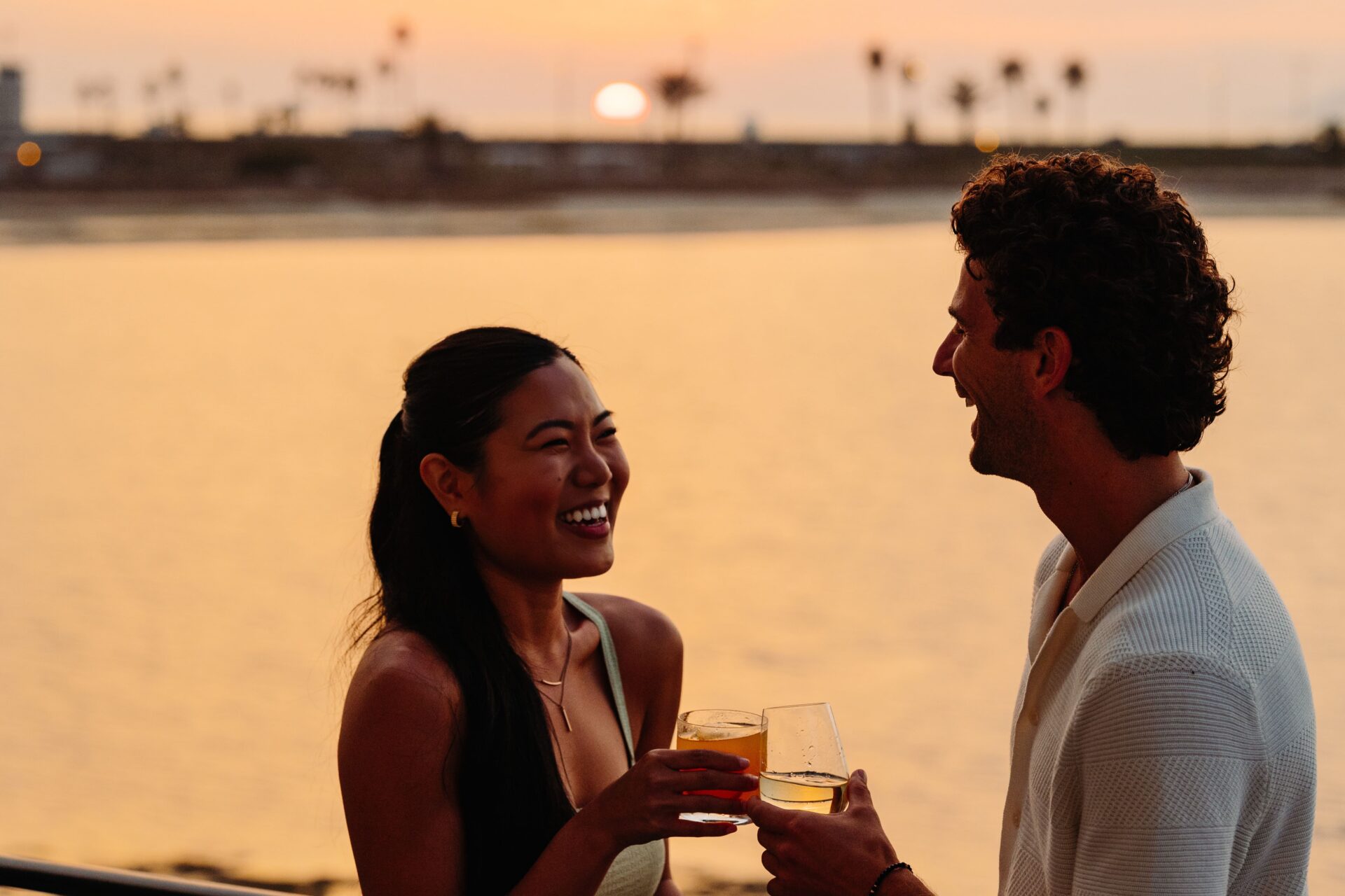 A woman and man stand by the water at sunset, smiling and holding drinks. The sky is orange and palm trees are visible in the background, creating a warm, relaxed atmosphere.