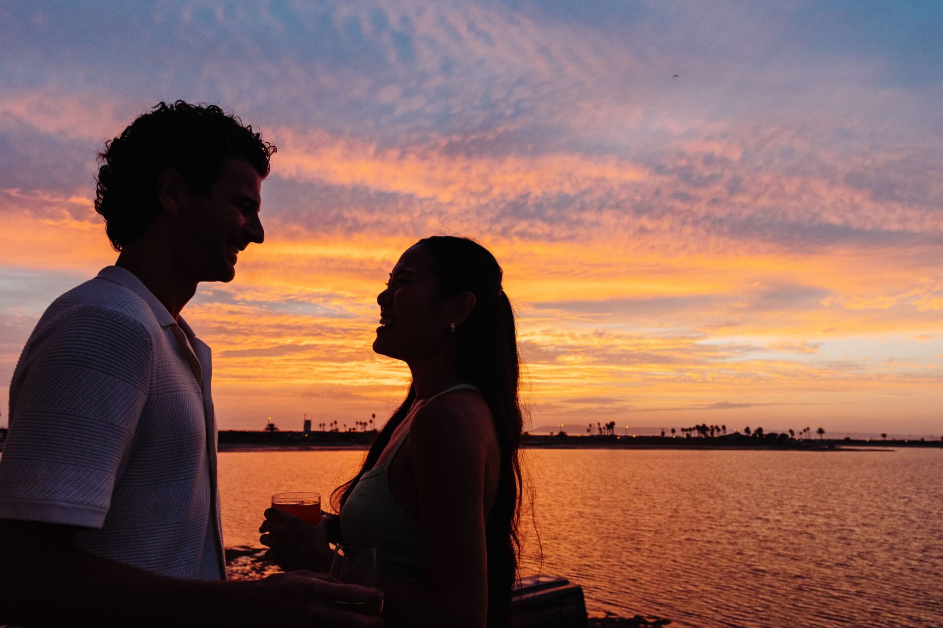 A couple stands by the water at sunset, facing each other and smiling. The sky is filled with vibrant orange, yellow, and purple hues, with silhouettes of palm trees in the distance.