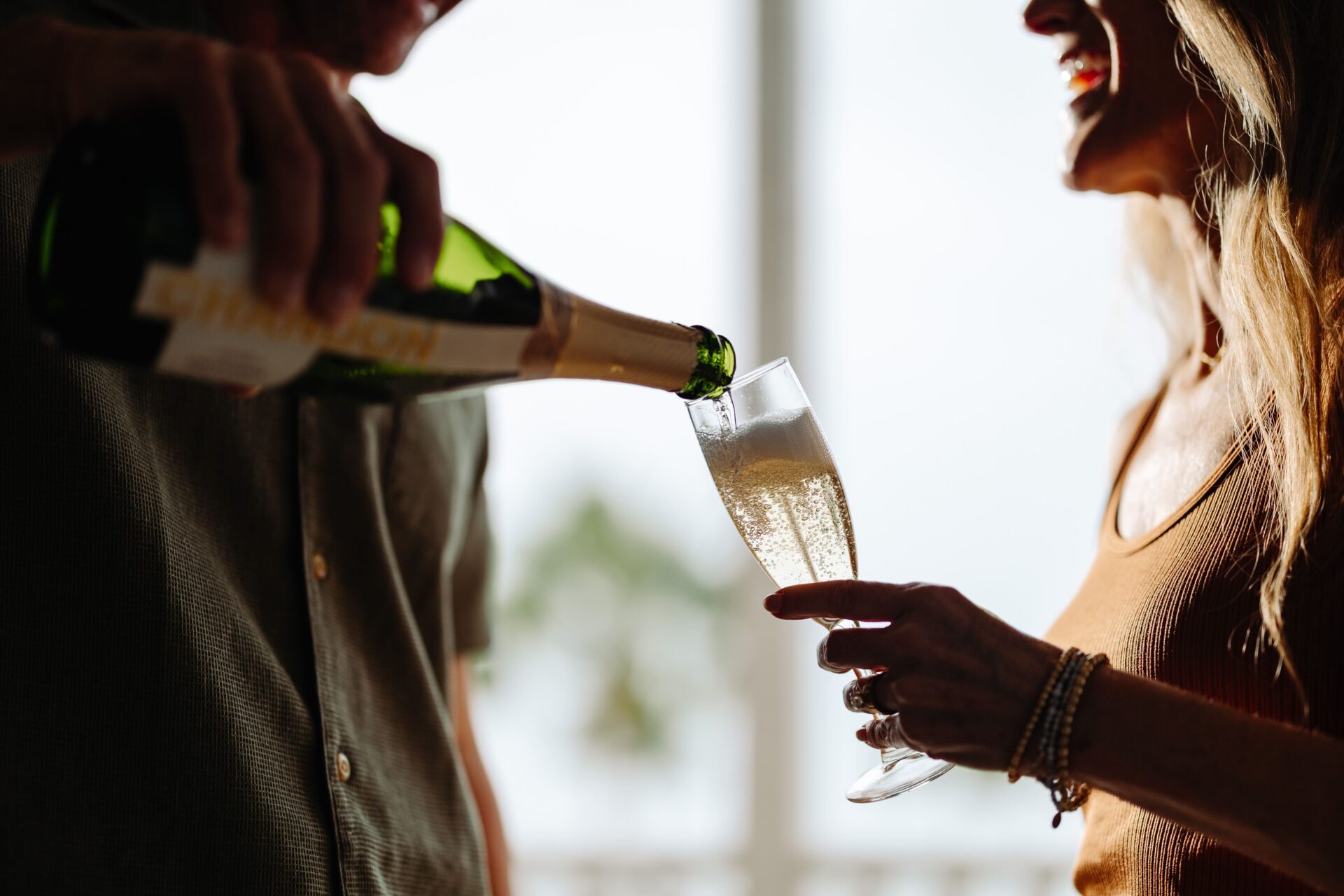 A person pours sparkling wine into a champagne flute held by another smiling person, both enjoying a bright, celebratory moment indoors.