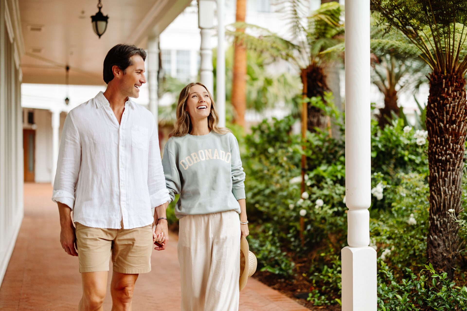 A man and woman walk hand in hand along a covered outdoor walkway lined with greenery. The man wears a white shirt and shorts; the woman wears a light sweatshirt and wide pants, smiling and holding a hat.