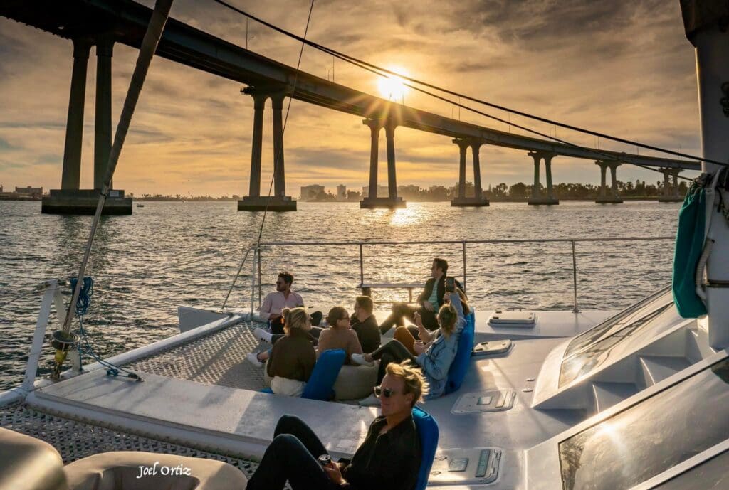 group of people on a boat under the coronado bridge with the sun setting in the background