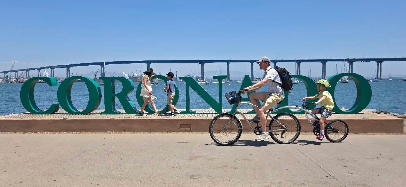 A man and a child ride bicycles past large “CORONADO” letters by the water, with a bridge and people walking in the background under a sunny sky.