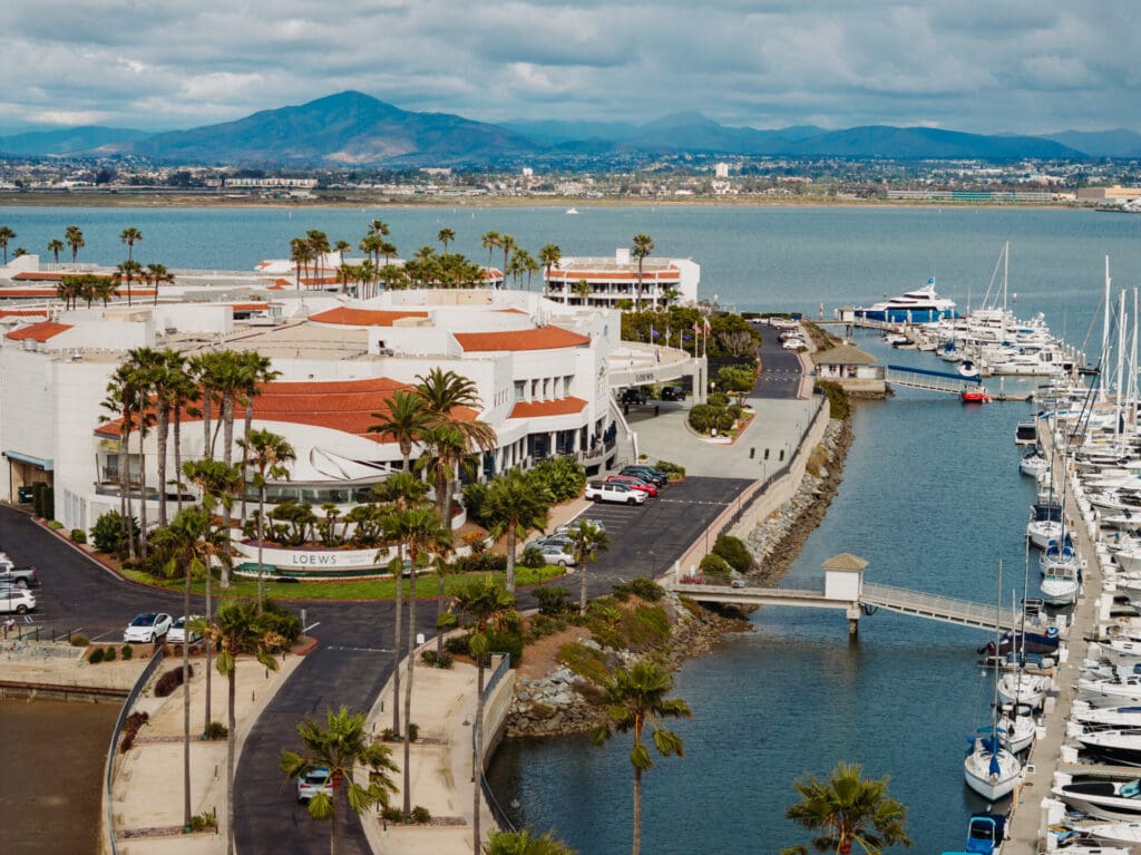 Aerial view of a marina with yachts docked next to a white resort-style building, palm trees, and a bay. Mountains and a cityscape are visible in the background under a cloudy sky.