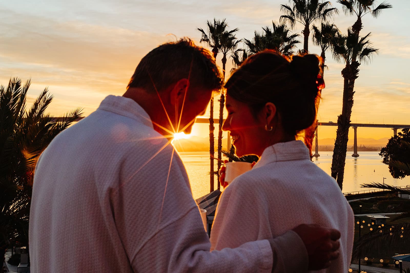 A couple in white robes stands close together at sunset, with the sun shining between them and palm trees in the background, creating a warm, romantic atmosphere by the water.