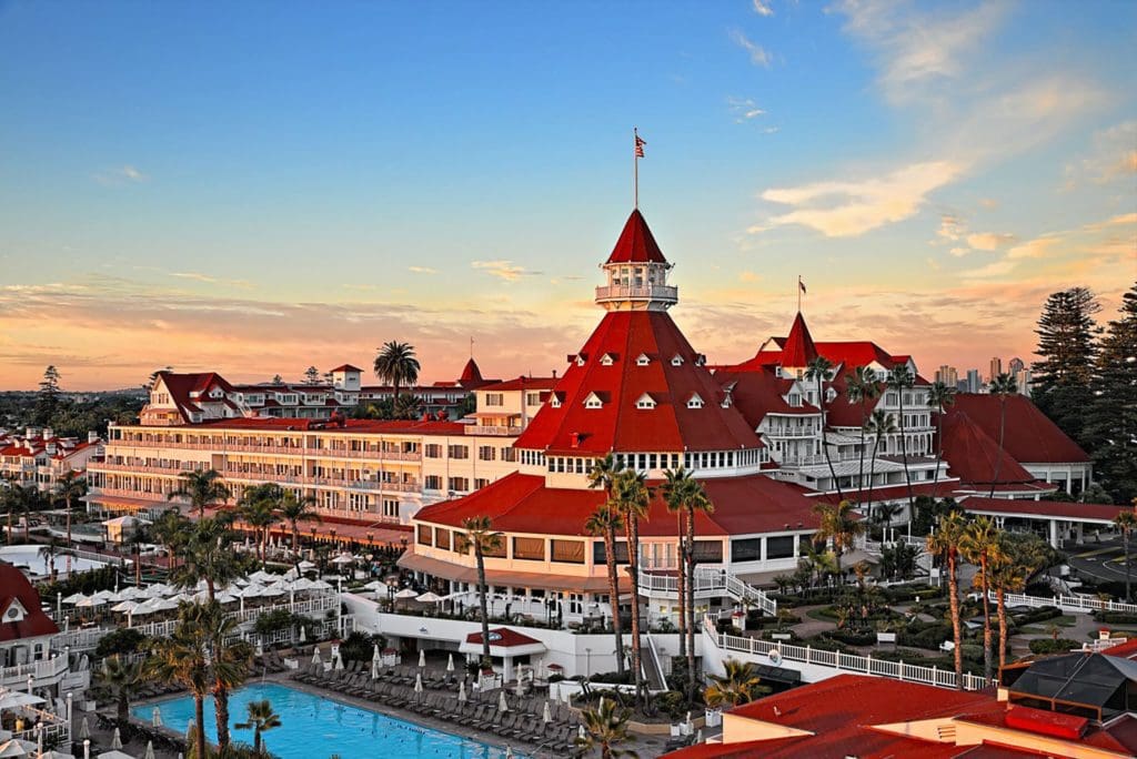 A large historic hotel with distinctive red roofs and white walls sits near a palm tree-lined pool at sunset, with a clear sky and city skyline in the background.