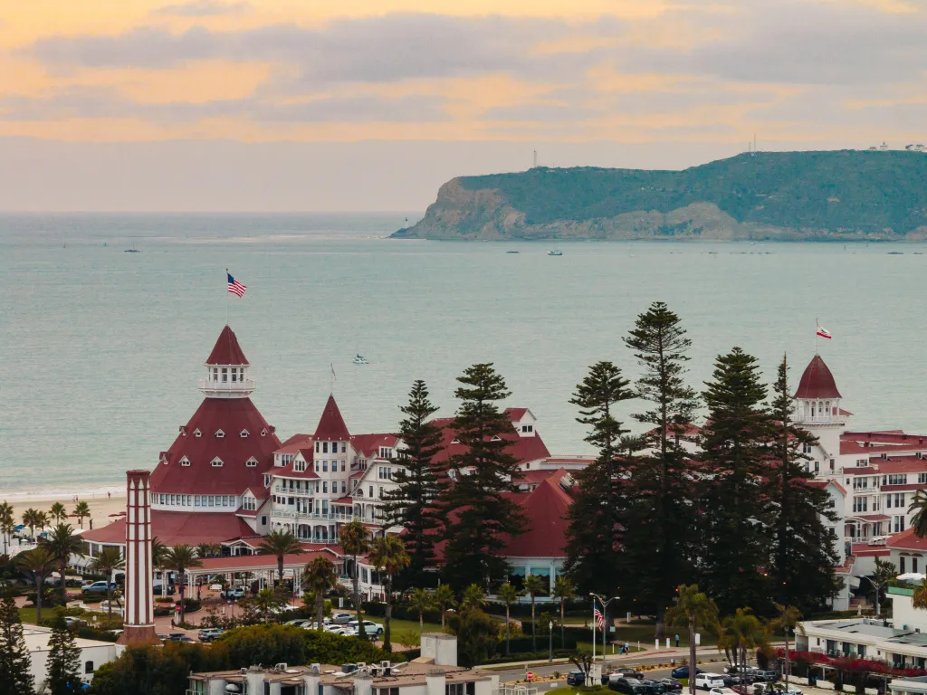 A historic beachfront hotel with red roofs and white walls sits by the ocean, bordered by tall trees, with cliffs and calm water in the background under a cloudy sky at sunset.