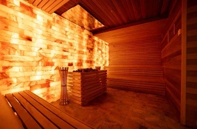 A warm, glowing sauna room with wooden benches, walls made of illuminated salt blocks, and a metal basket filled with sauna stones in the center.