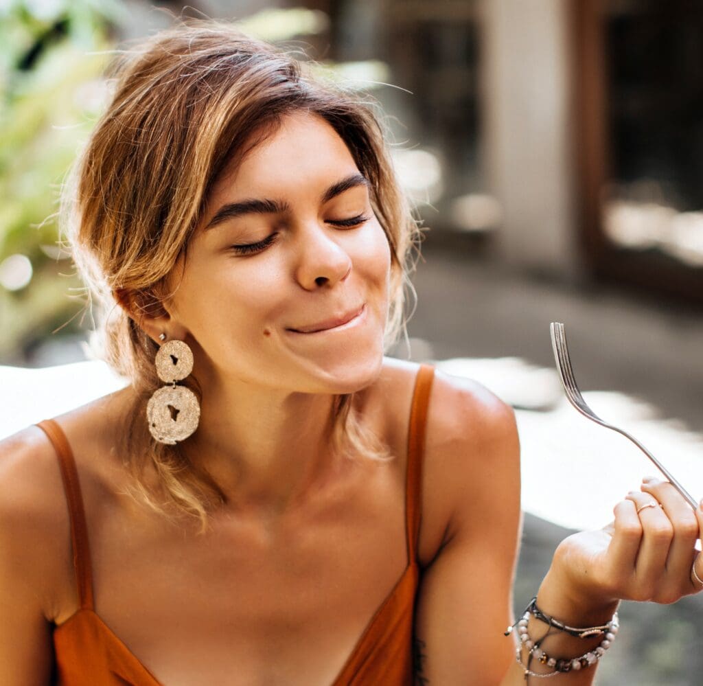 A woman with short hair and large earrings smiles with her eyes closed, appearing to savor food. She holds a fork in one hand and wears a sleeveless top, sitting outdoors in natural light.